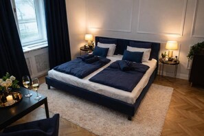 Ein Schlafzimmer mit einem großen Bett, zwei Nachttischen, einem Beistelltisch, einem Fenster mit Vorhängen und einem kleinen Tisch mit einer Vase und Gläsern.