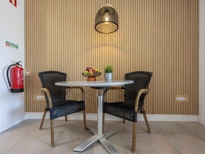Ein Zimmer mit einem runden Tisch, zwei Korbstühlen, einer Topfpflanze, einer Obstschale, einem Feuerlöscher und einer an der Wand befestigten Leuchte.