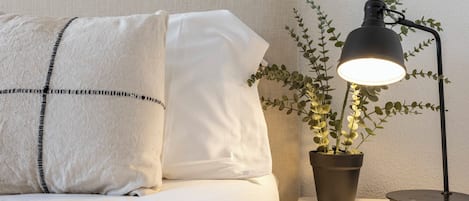 A bed with white bedding and two pillows, a small potted plant with green leaves, and a black desk lamp on a bedside table.