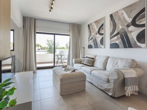 Un salon moderne avec un canapé, un pouf et une table à manger. Il y a un balcon avec vue sur l’extérieur.