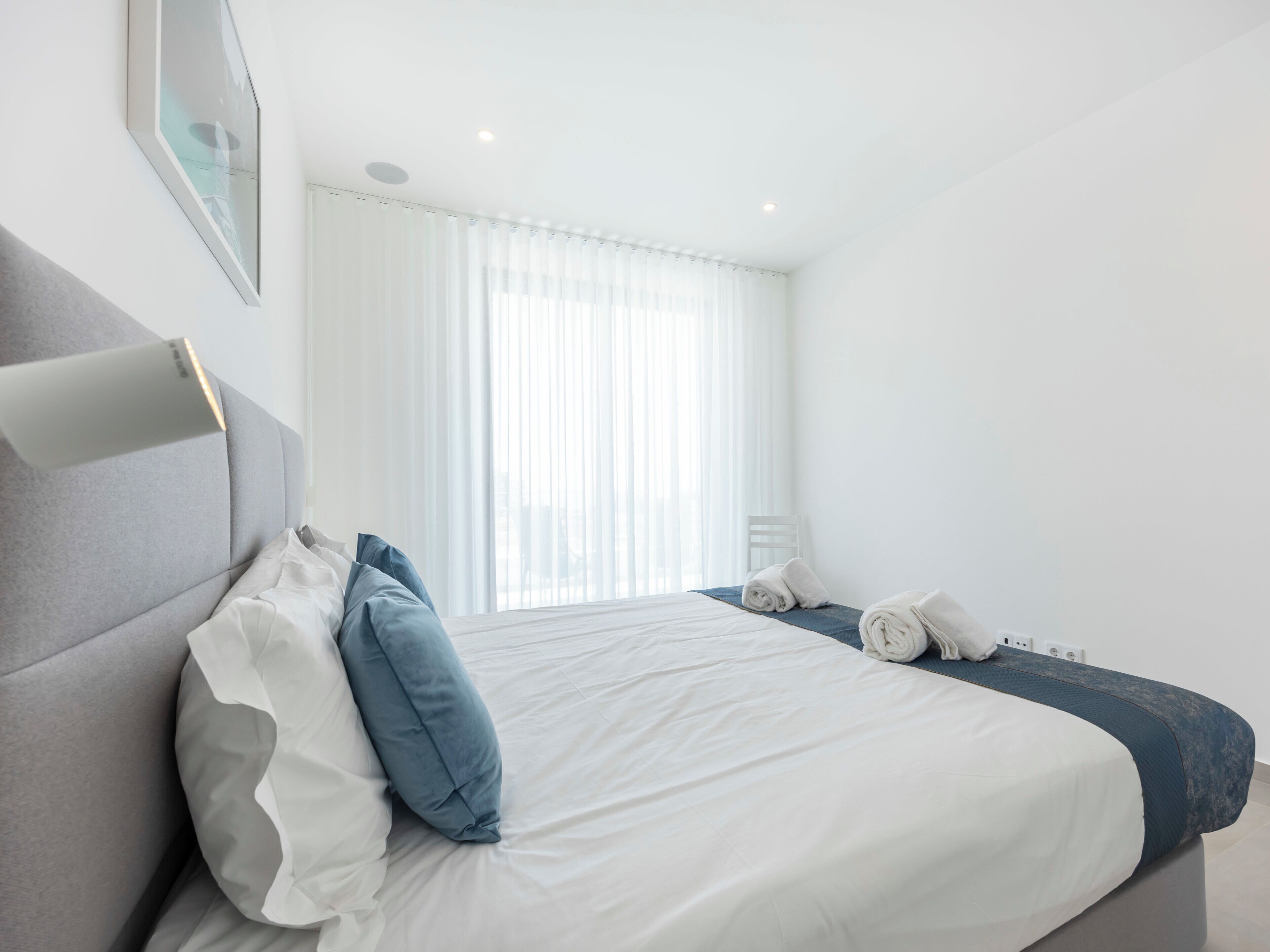 Une chambre d’hôtel moderne avec un grand lit, une table de chevet, une chaise et une fenêtre avec des rideaux légers.