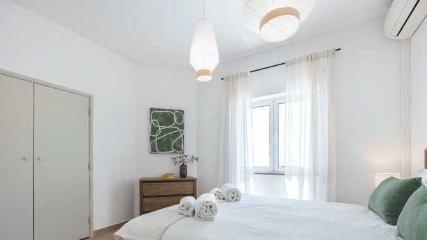 A bedroom with a bed, a bedside table with a vase, a window with curtains, and a wall-mounted artwork. - Casa Horizonte - Old Town Lagos (Lagos)