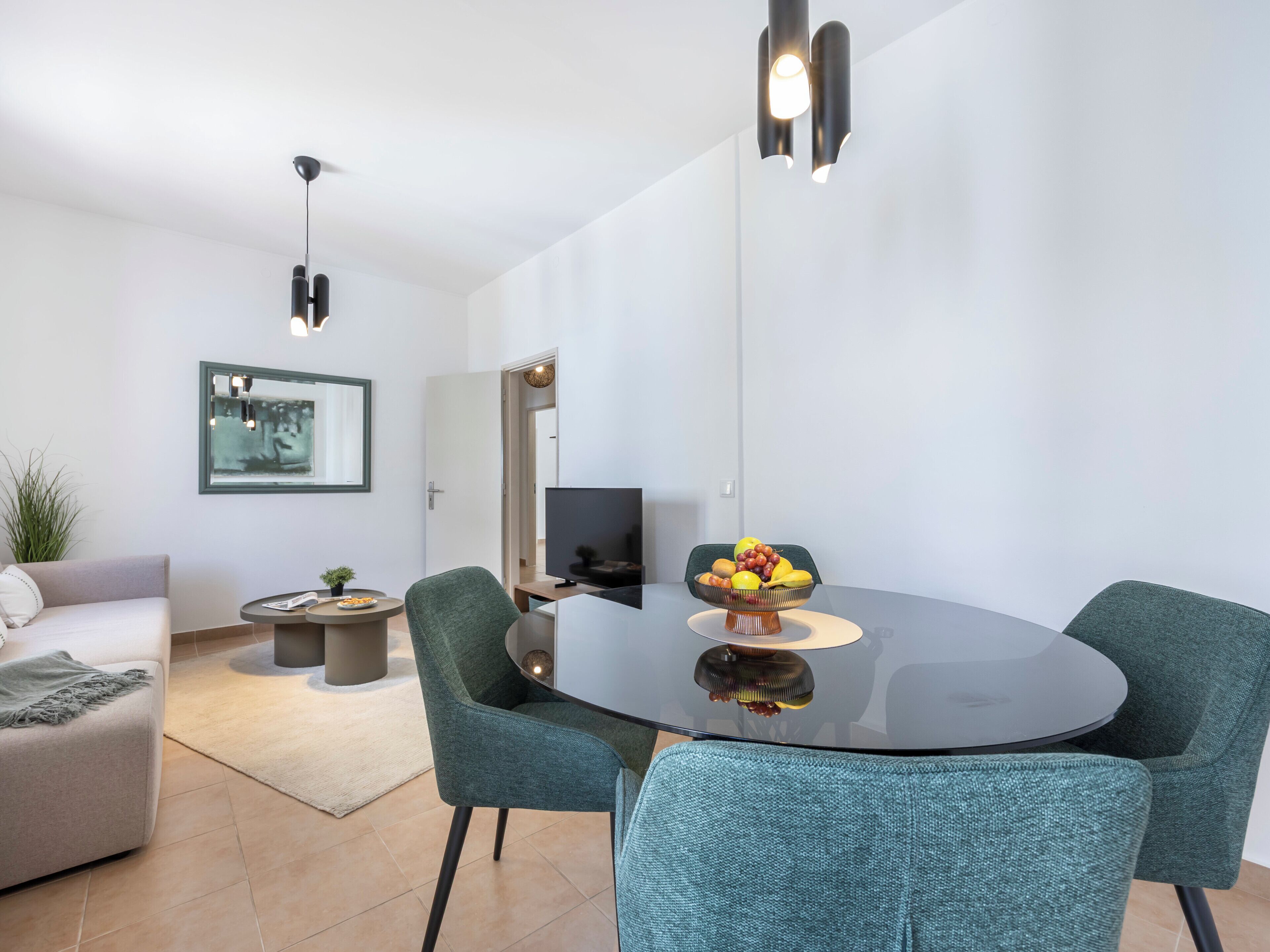 A modern living room with a round glass table, teal chairs, a flat-screen TV, and a fruit bowl on the table.