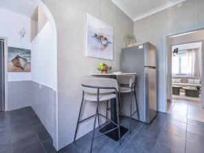 A modern kitchen with a refrigerator, two bar stools, a small table with a bowl of fruit, and a painting on the wall. - Casa Horizonte - Old Town Lagos (Lagos)