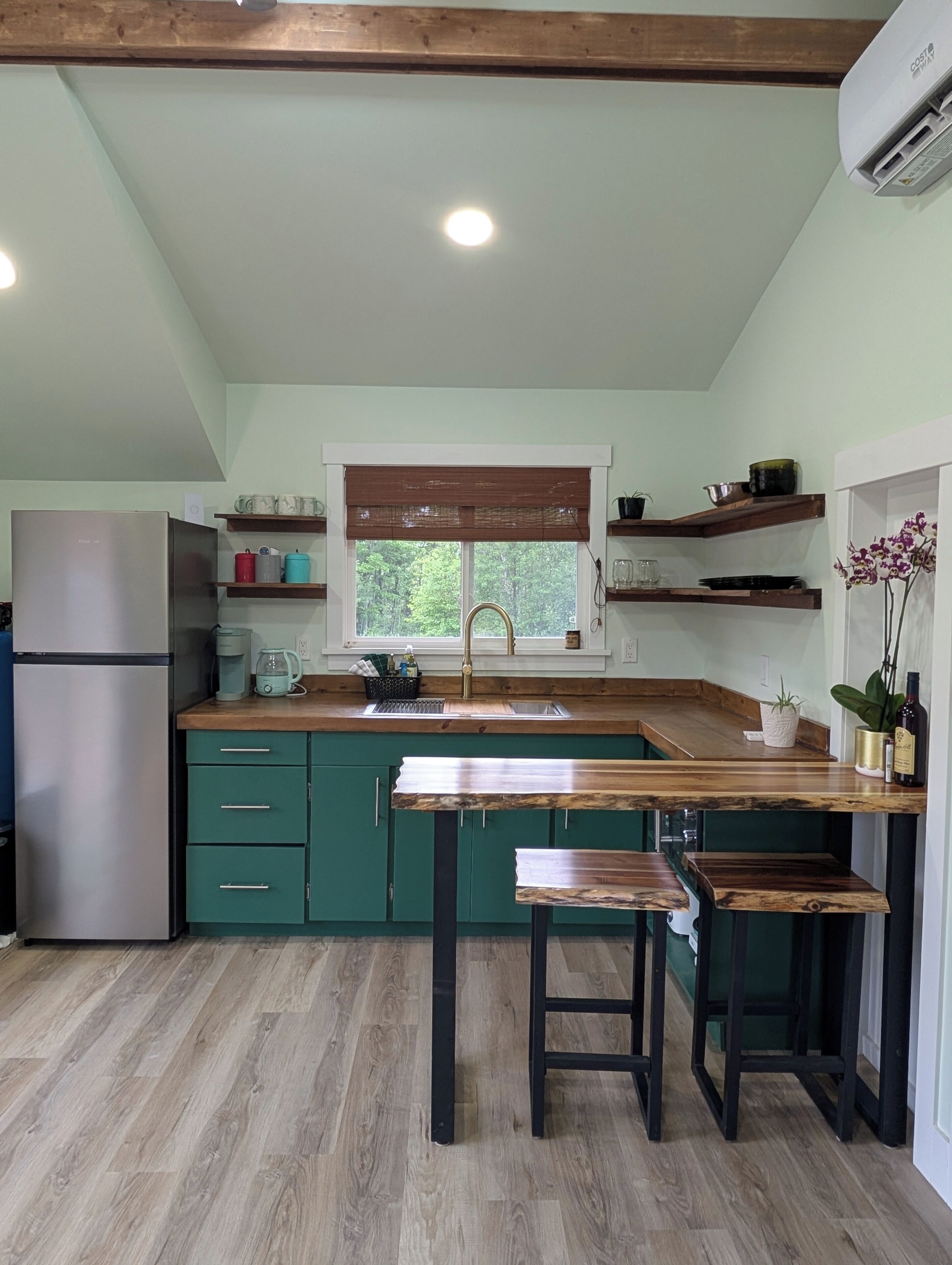 Une cuisine équipée d’un réfrigérateur encastré, d’un plan de travail en bois, de meubles de rangement verts et d’une fenêtre donnant sur un paysage verdoyant.