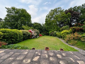 Um jardim com um caminho pavimentado que leva a um gramado, cercado por arbustos e árvores diversos.