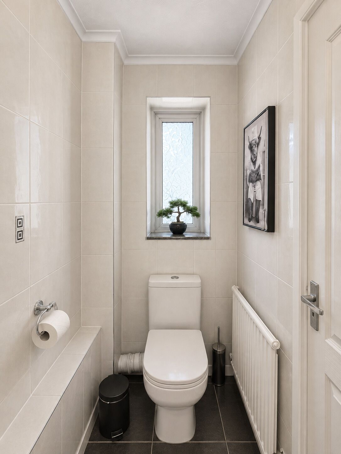 A bathroom with a toilet, a bidet, a trash bin, a radiator, and a window with a plant.