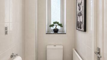 A bathroom with a toilet, a bidet, a trash bin, a radiator, and a window with a plant.