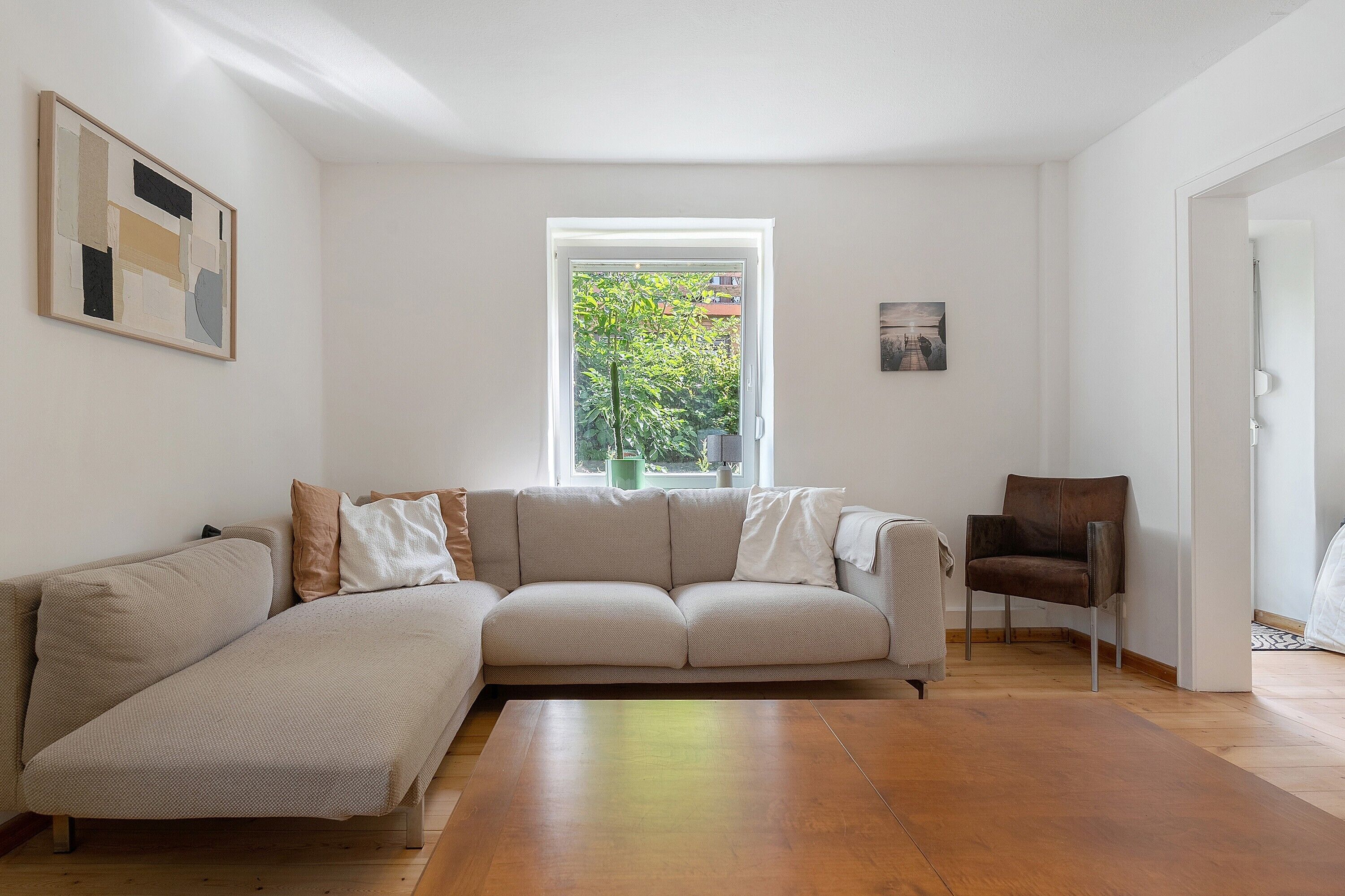 A living room with a sectional sofa, a single chair, a window with a view of greenery, and a painting on the wall.