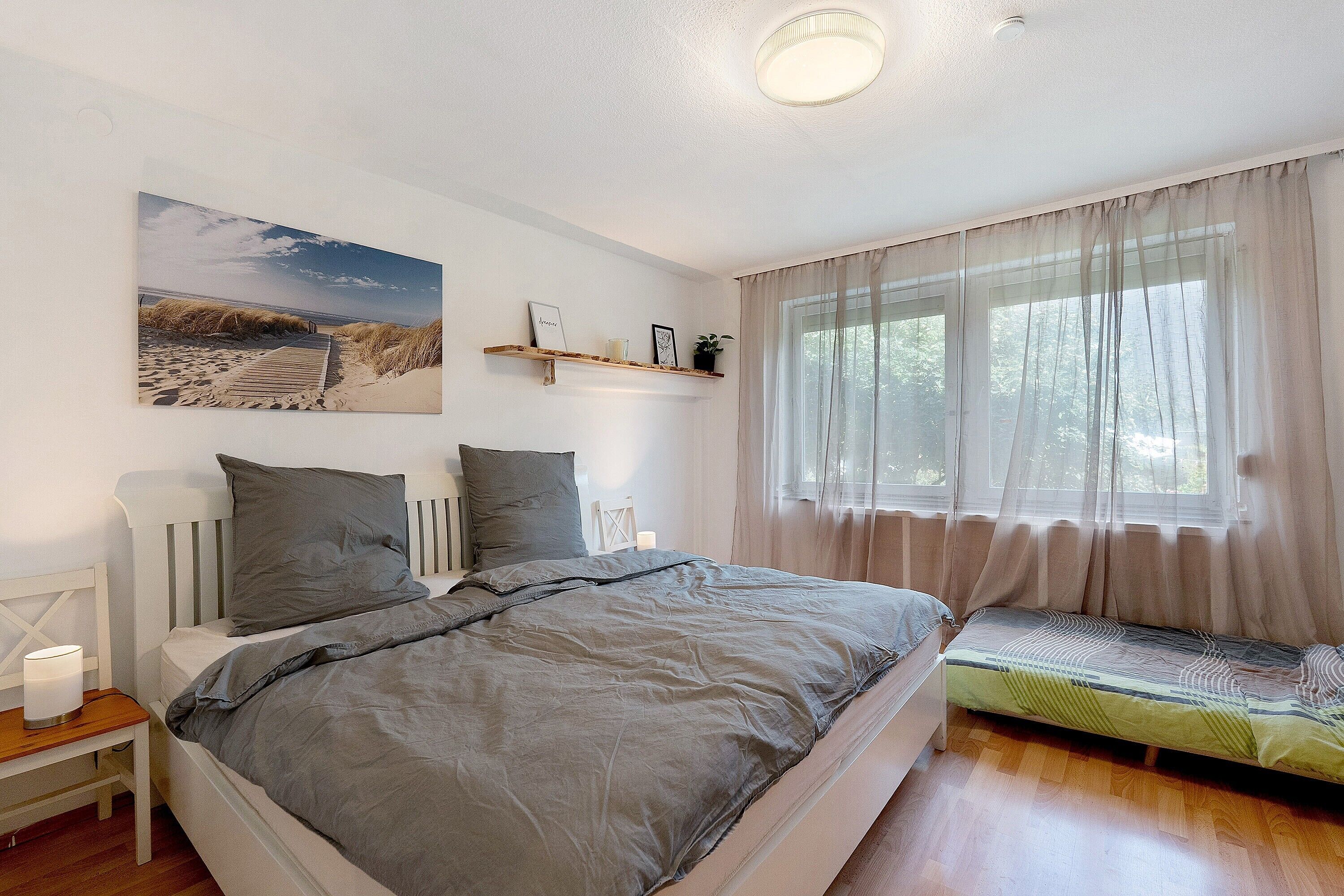 Een slaapkamer met een groot bed, nachtkastjes, een raam met dunne gordijnen en een wandplank met decoratieve spullen.