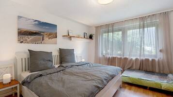 Een slaapkamer met een groot bed, nachtkastjes, een raam met dunne gordijnen en een wandplank met decoratieve spullen.