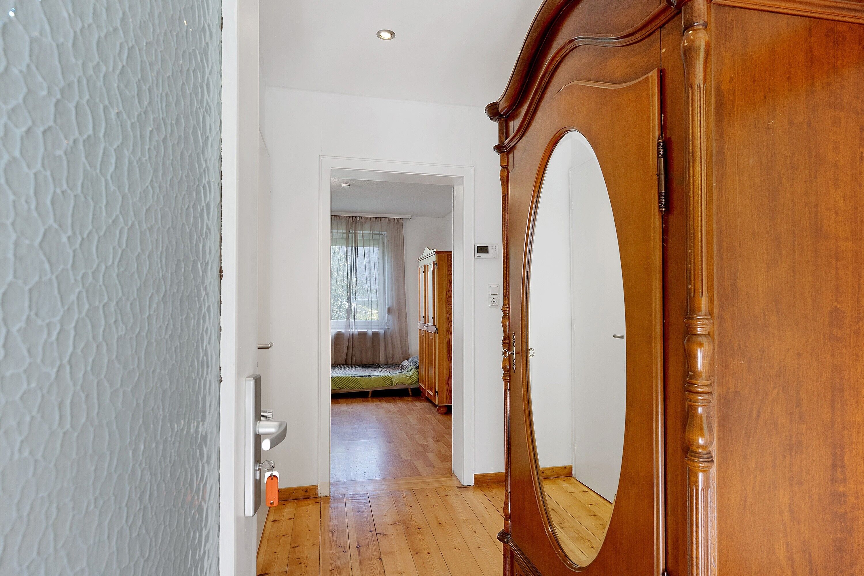Un couloir avec une porte en bois, un miroir et un lit, avec une fenêtre visible au fond.