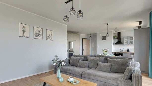 A modern living room with a grey sofa, a wooden coffee table, and a grey rug. There are framed artworks on the wall, a kitchen in the background, and pendant lights above the sofa. - Letnica 159 | Sea View | 2 Bedrooms (Gdańsk)