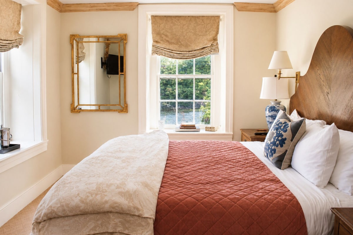 A bedroom with a large bed, a window with a view of greenery, a mirror, and a bedside table with a lamp.