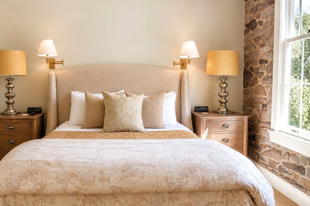 A bedroom with a large bed, two bedside tables with lamps, a stone wall, and a window.