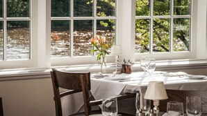 A dining table set for two with a view of a body of water through large windows. - Golden Pheasant Inn (Erwinna)