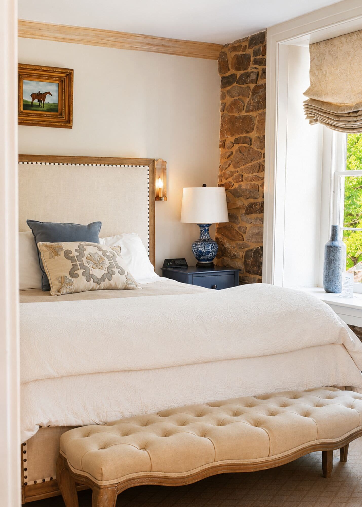 Un dormitorio con una cama grande, un banco, una mesita de noche con una lámpara, un cuadro en la pared y una ventana con vistas.