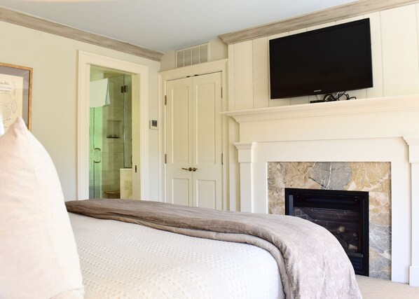 A bedroom with a bed, a television mounted above a fireplace, and a door leading to a bathroom.