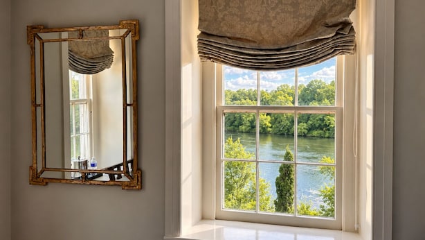A room with a large window overlooking a river, a mirror with an ornate frame, and a window treatment with a floral pattern. - Golden Pheasant Inn (Erwinna)