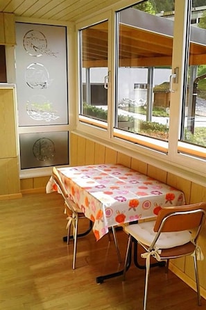 A room with a wooden floor, a table with a floral tablecloth, two chairs, and a window with a view of the outside. - Bergtreu 13 # 2-bed apartment, Dusche, WC 2-Bettwohnung, Dusche, WC by Interhome (Leukerbad)
