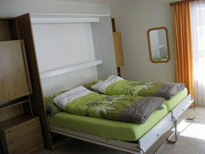 A double bed with green bedding and two pillows, a wooden wardrobe, a mirror, and a window with curtains. - Bergtreu 13 # 2-bed apartment, Dusche, WC 2-Bettwohnung, Dusche, WC by Interhome (Leukerbad)