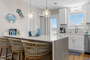 A modern kitchen with a central island, bar stools, and decorative wall art. - Infinity Dream𓇼Ocean Views+Sleeps 14+Golf Cart (Oak Island)