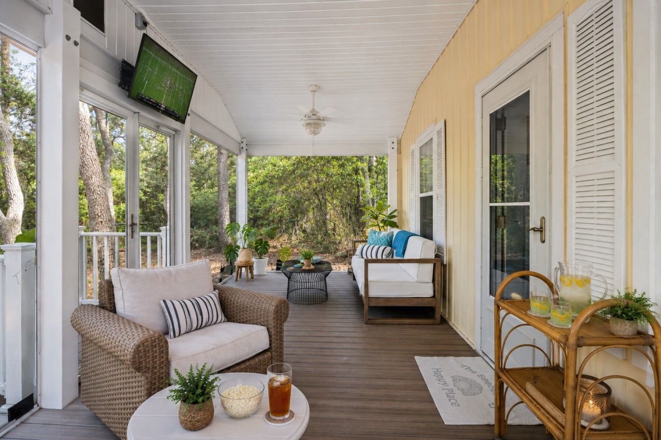 Eine überdachte Veranda mit einer Couch, Stühlen, einem Tisch mit Getränken und einem an der Wand befestigten Fernseher.