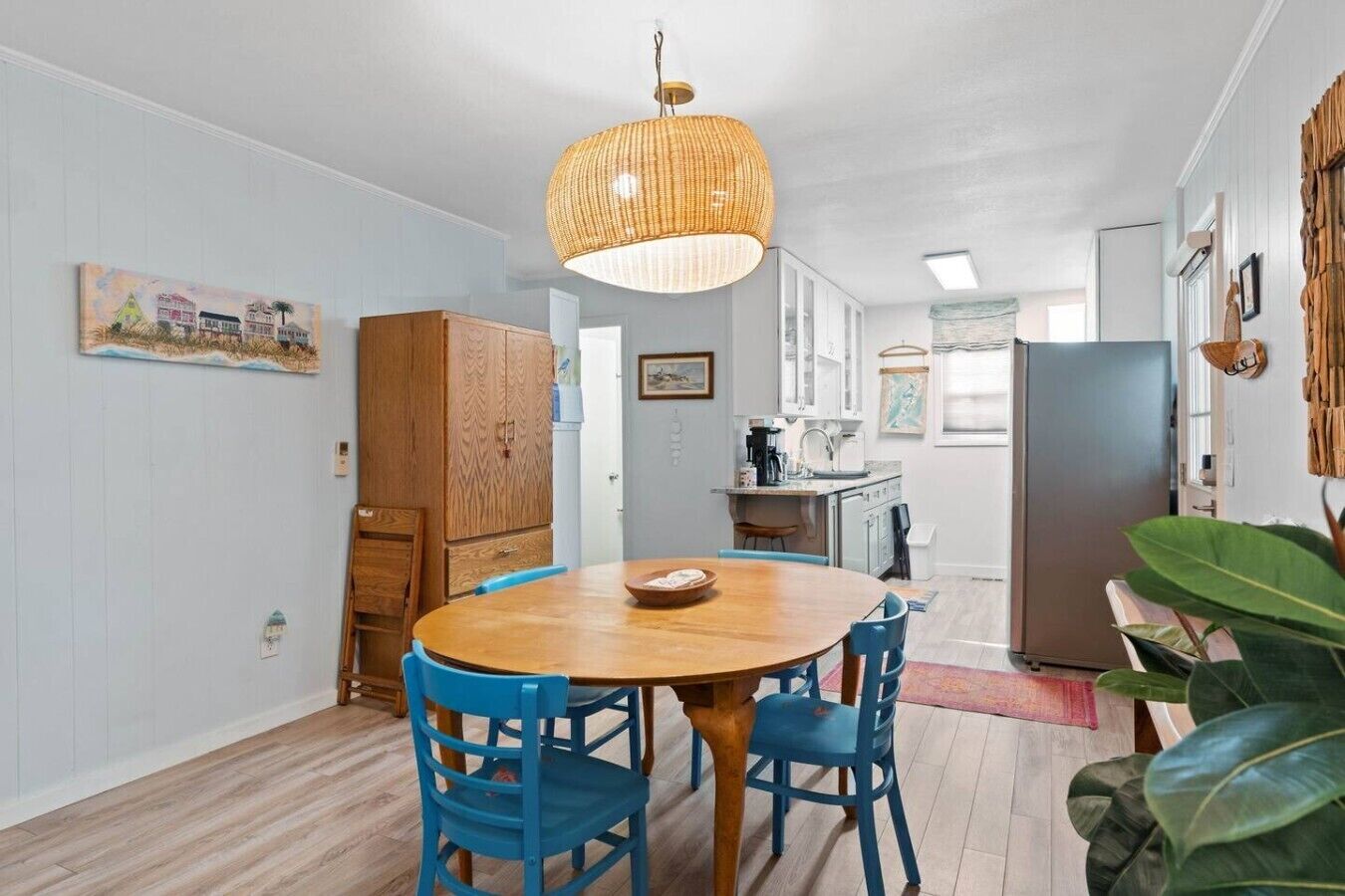 A dining area with a round wooden table and blue chairs, a hanging wicker light fixture, and a kitchen in the background.