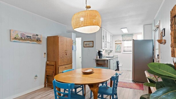 A dining area with a round wooden table and blue chairs, a hanging wicker light fixture, and a kitchen in the background. - Birdsong Cottage𓇼Island Retreat+Central Location (Oak Island)