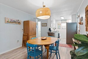 A dining area with a round wooden table and blue chairs, a hanging wicker light fixture, and a kitchen in the background. - Birdsong Cottage𓇼Island Retreat+Central Location (Oak Island)