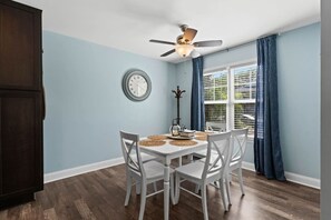 A dining area with a table set for four, a ceiling fan, a clock, and a window with blinds. - My Three Buoys𓇼Sleeps 10+Game Room+Spacious (Oak Island)