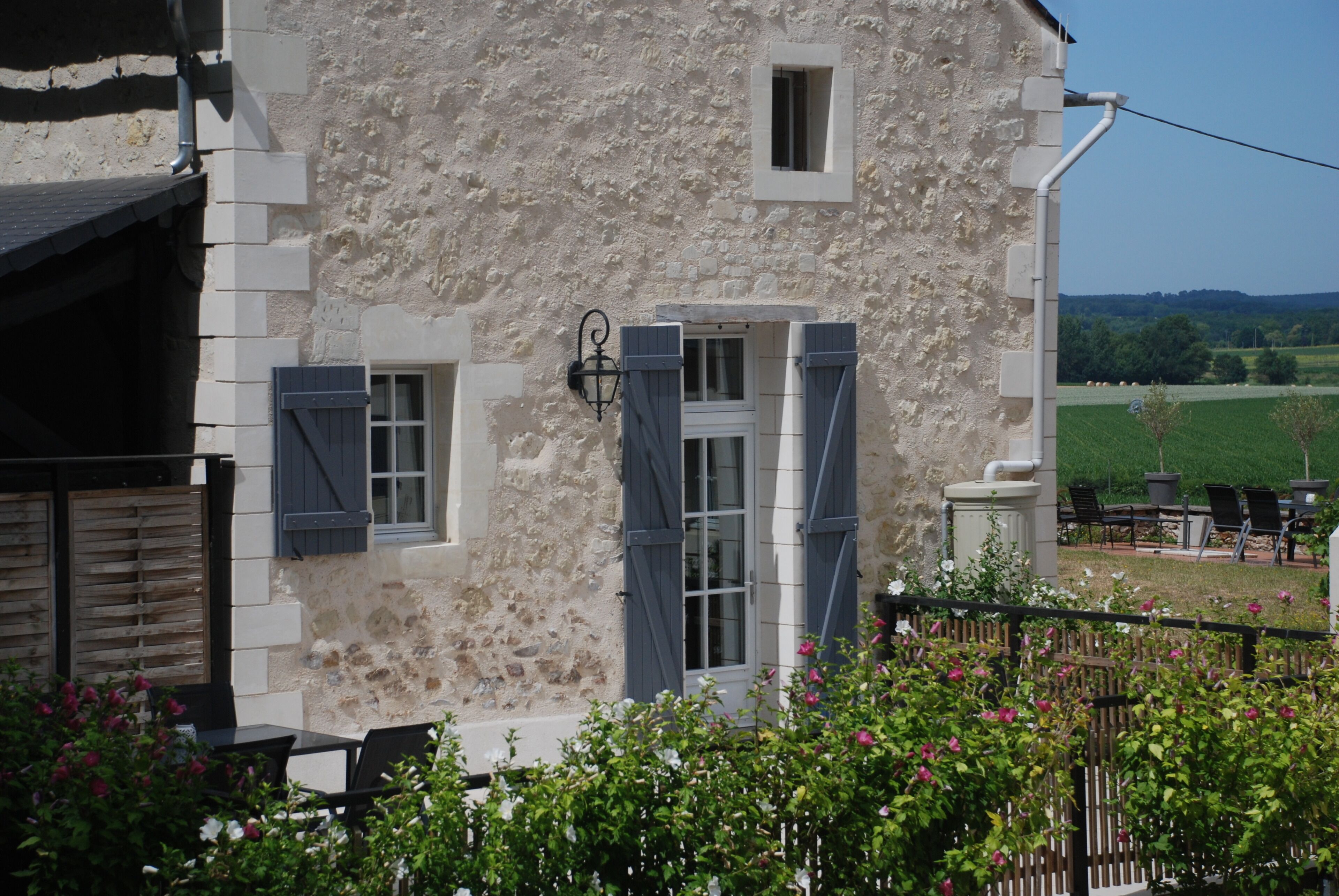 A stone building with a window and shutters, surrounded by greenery and a fence.
