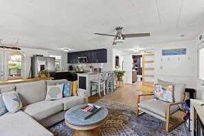 A spacious living room with a sofa, armchair, and coffee table, a kitchen with dark cabinets and a dining area with a table and chairs. - Sandy Swan𓇼Cozy Cottage Vibes+Golf Cart+Hot Tub (Oak Island)