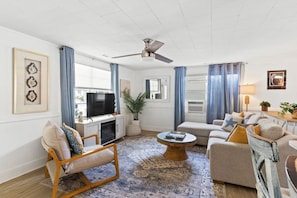 A living room with a television, seating area, and a ceiling fan. - Sandy Swan𓇼Cozy Cottage Vibes+Golf Cart+Hot Tub (Oak Island)