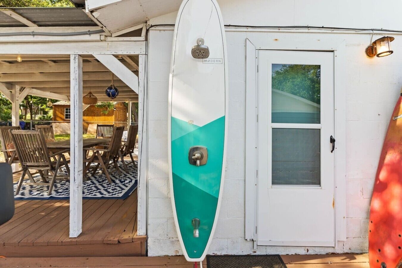 A white building with a door, a covered patio area with wooden furniture, and a surfboard leaning against the building.