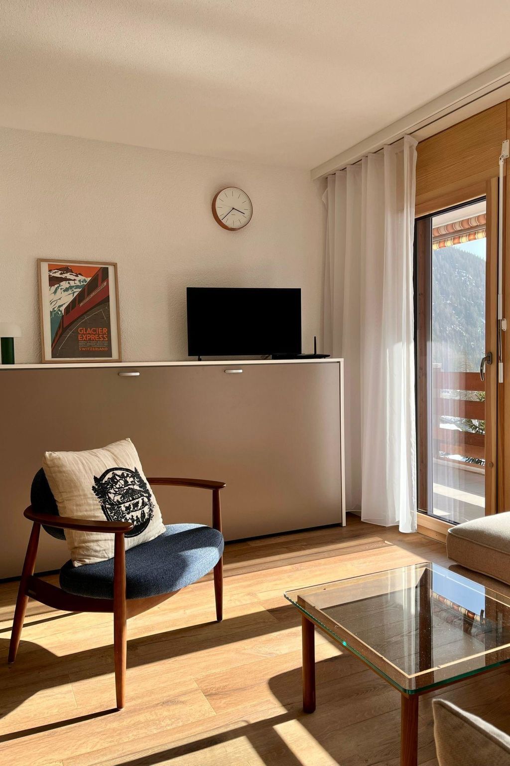 Une pièce avec un plancher en bois, une table en verre, une chaise avec un coussin, un téléviseur à écran plat posé sur un meuble, une horloge au mur et une fenêtre avec des rideaux.
