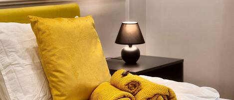 A bed with white bedding, a yellow pillow, and rolled towels.