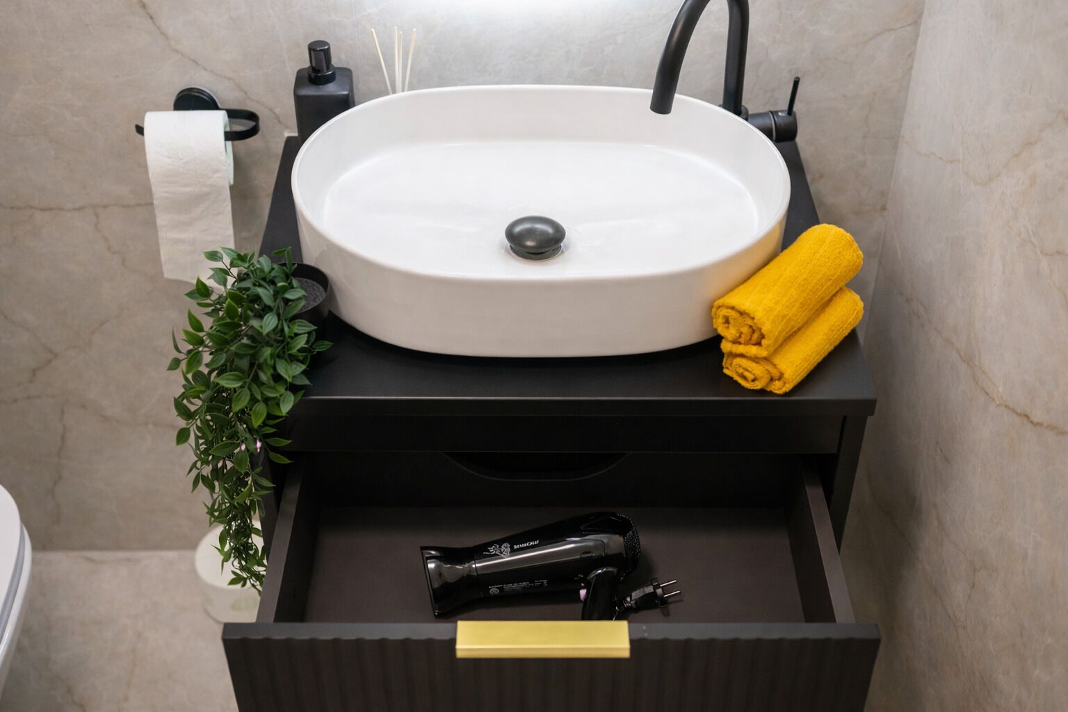 A bathroom with a white sink, black faucet, and a black countertop. There is a yellow towel and a green plant on the counter.