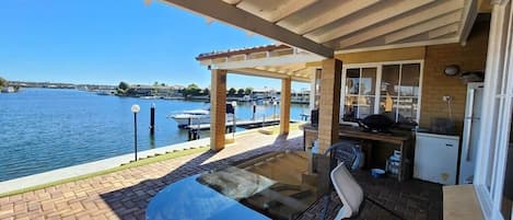 En overdækket terrasse med et bord og stole, udsigt til vandet og et lille tekøkken.