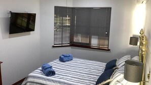 Ein Schlafzimmer mit einem Bett, einem an der Wand montierten Fernseher, einem Fenster mit Jalousien, einem Deckenventilator und einer Nachttischlampe.
