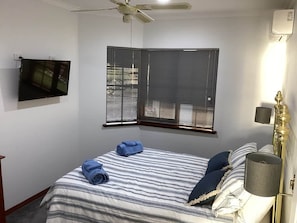 A bedroom with a bed, a television mounted on the wall, a window with blinds, a ceiling fan, and a bedside lamp. - Mandurah Holiday Apartments ‘Pelican’ (Dudley Park)