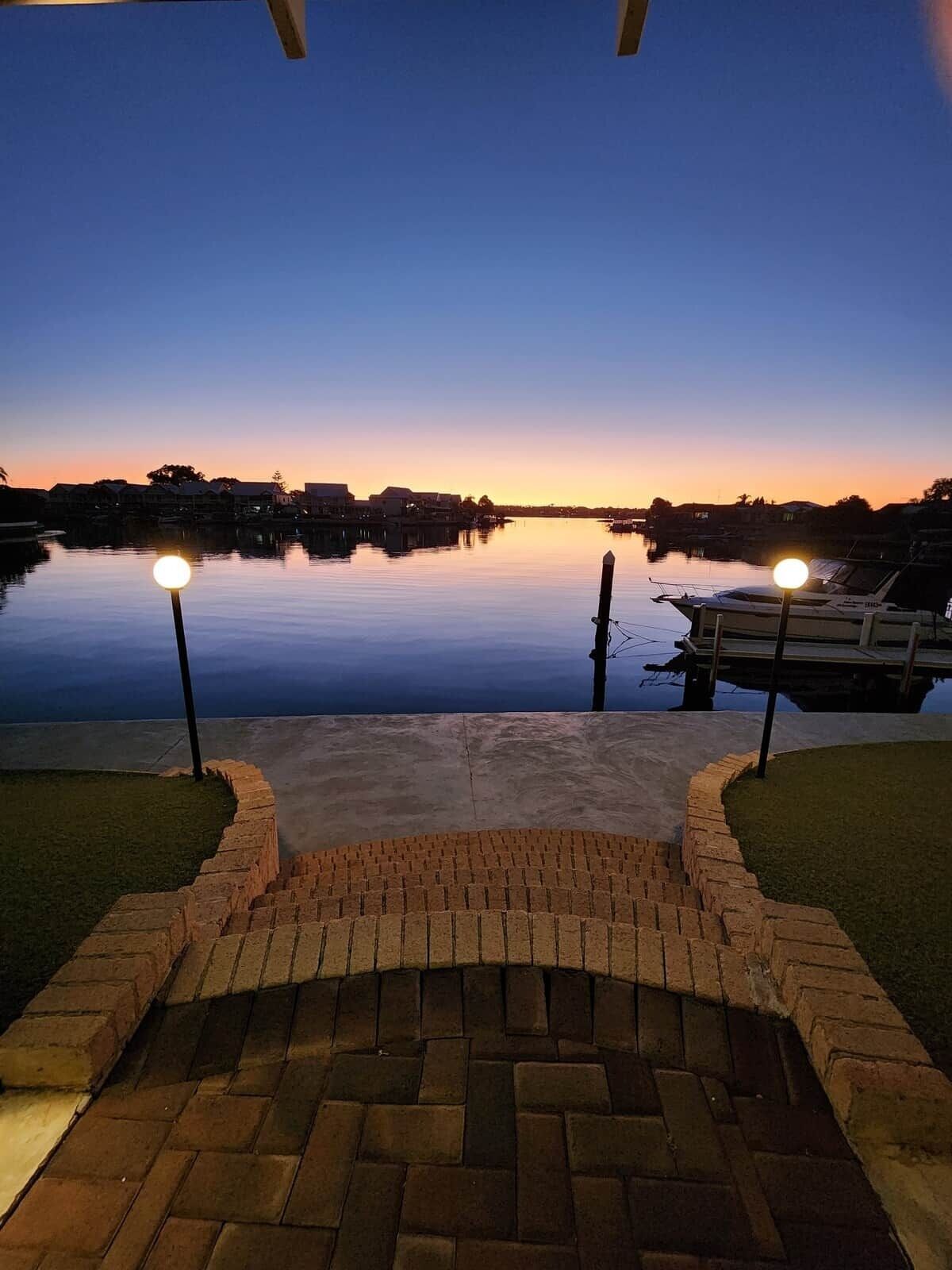 A waterfront patio with a brick pathway leading to a dock area, surrounded by calm water and a sunset sky.