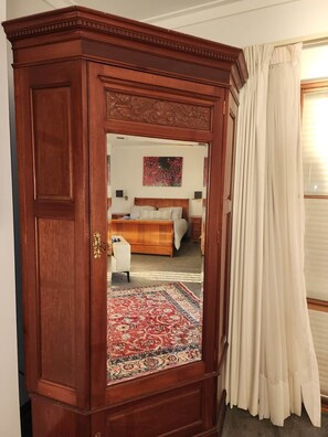Une chambre à coucher avec une armoire en bois, un lit avec une tête de lit, une table de chevet et une commode, ainsi qu’une fenêtre avec des rideaux.