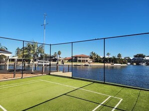 A fenced tennis court with a water body in the background. - Mandurah Holiday Apartments ‘Osprey’ (City of Mandurah)