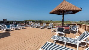 Een houten terras met gestreepte ligbedden en een centrale tafel, omgeven door een houten omheining met uitzicht op een strand.