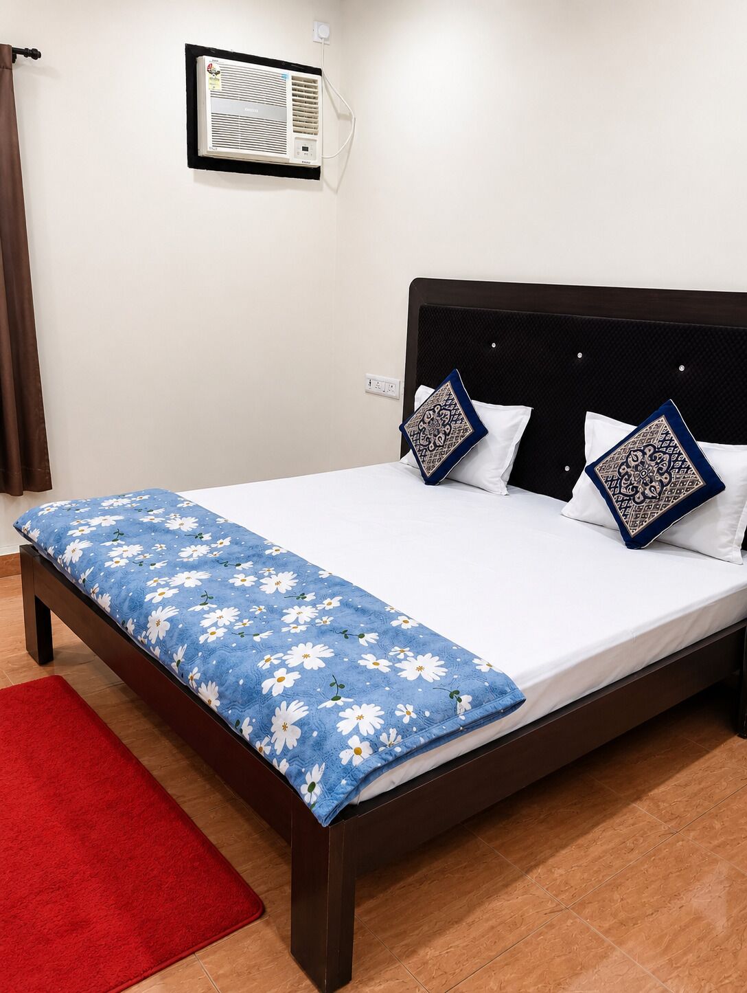 Un letto con una coperta azzurra a motivi floreali, due cuscini decorati, un tappeto rosso e un condizionatore d'aria fissato al muro.