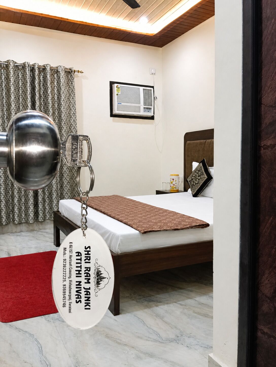 A hotel room with a bed, a bedside table, a lamp, a window with curtains, and a wall-mounted air conditioning unit.