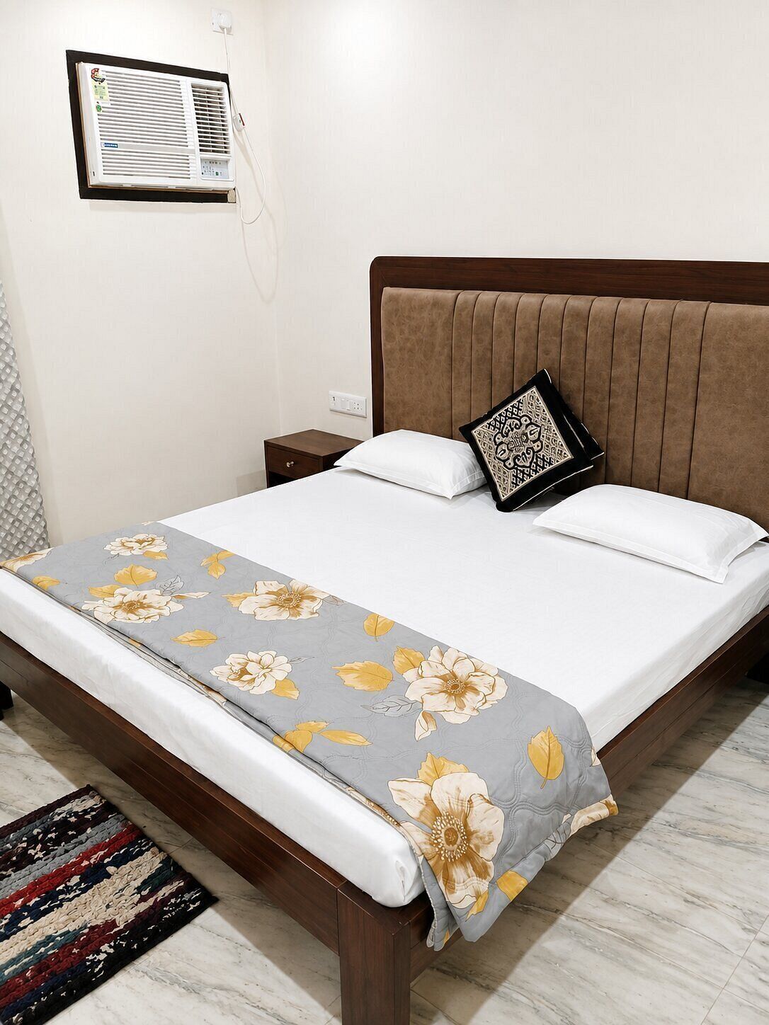 A hotel room with a bed, a nightstand, a wall-mounted air conditioner, and a patterned rug.
