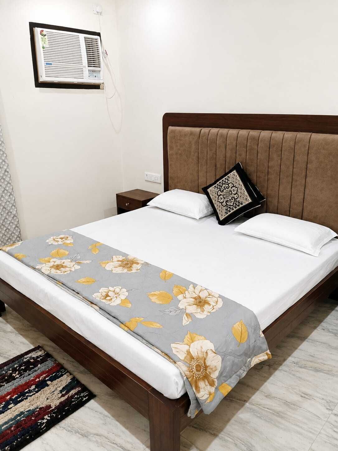A hotel room with a bed, a bedside table, a nightstand, a wall-mounted air conditioner, and a patterned rug.