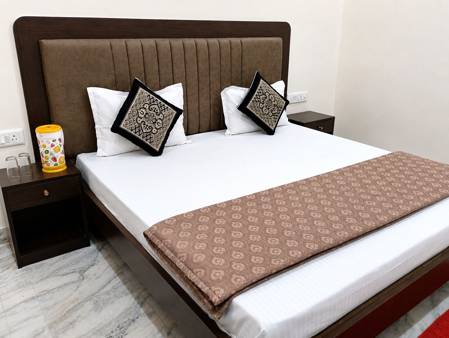 A hotel room with a large bed, two nightstands, a bedside table, a lamp, and a patterned throw blanket.